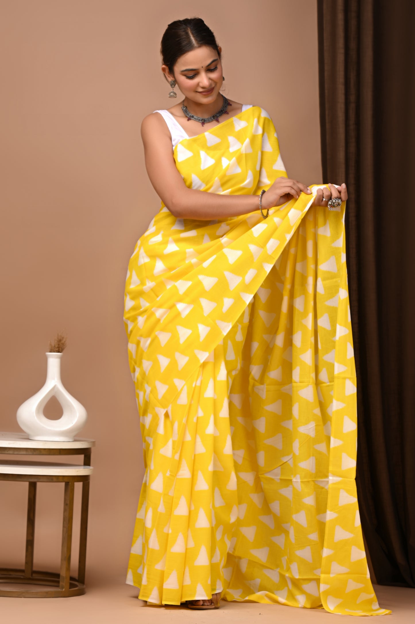 A woman models a bright yellow saree with a white geometric triangle pattern, paired with a white sleeveless blouse, standing in a studio setting with a beige backdrop and a small side table.